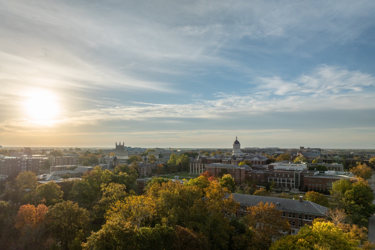 Aerial view of a cityscape at sunrise. The sun is low on the horizon, casting a warm glow over lush trees and historic buildings under a partially cloudy sky.
