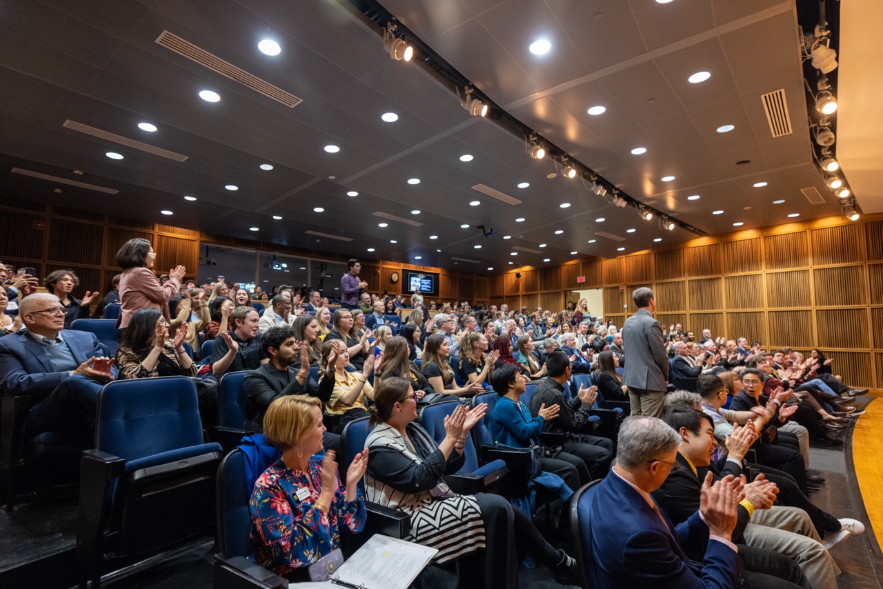 Audience in a modern lecture hall applauding a standing speaker. The room is full, lights are bright, and the mood is enthusiastic and engaged.