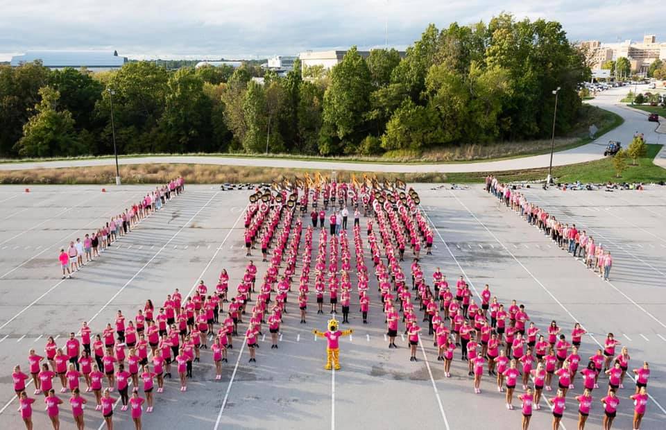 Members of Marching Mizzou wearing pink shirts, standing in a marching band formation forming a pink ribbon for breast cancer awareness
