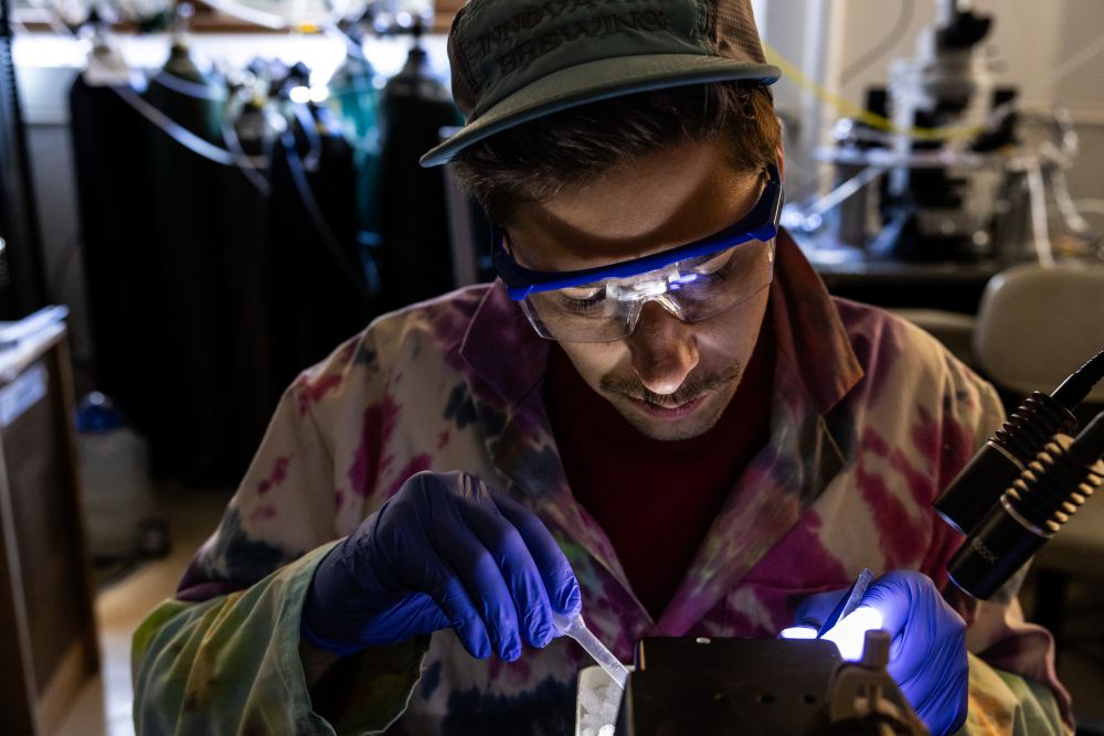 A scientist in a tie-dye lab coat works intently with tools. Wearing protective eyewear and gloves, he's illuminated by focused lights in a lab setting.