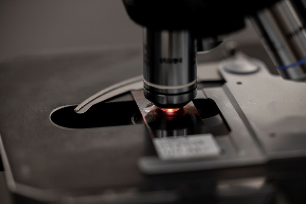 Close-up of a microscope lens focused on a glass slide with a reddish glow, suggesting an active sample analysis in a laboratory setting.