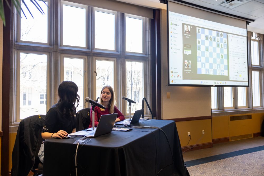 two women sit at table beside projection screen showing chess board