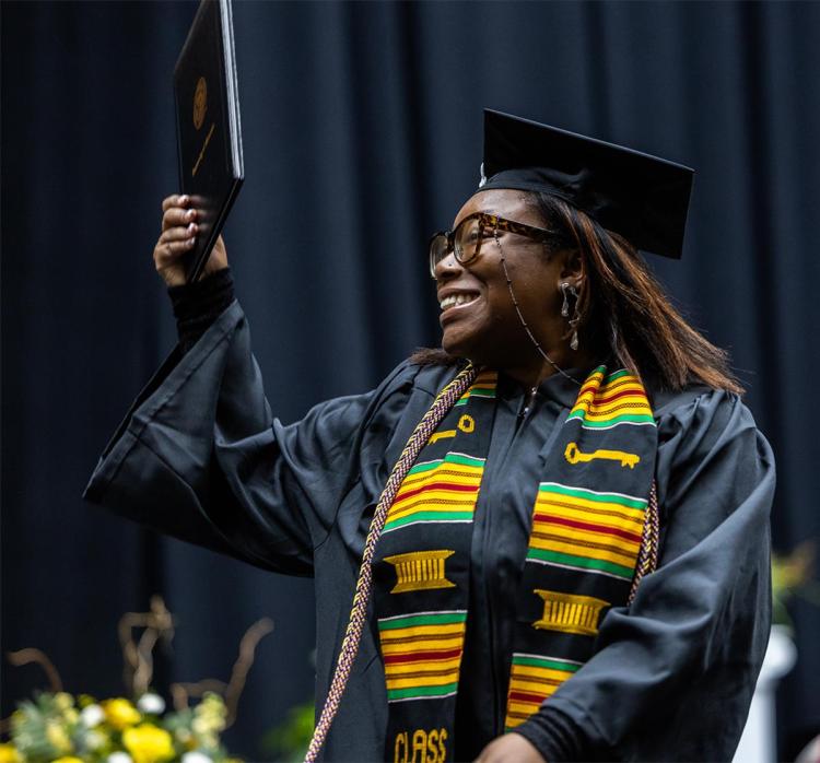 A student looking away from the camera, smiling while holding the diploma in their right hand in the air.