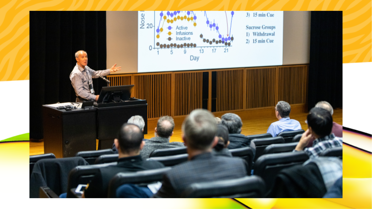 A speaker gestures towards a data graph on a large screen in a lecture hall. Engaged audience members are seated, with a focused, informative tone.