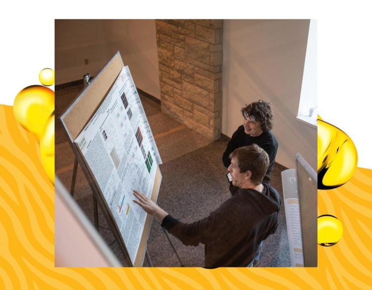 Two people examine a large, detailed poster on a stand in a well-lit room. The scene conveys focus and engagement. Abstract yellow patterns frame the image.