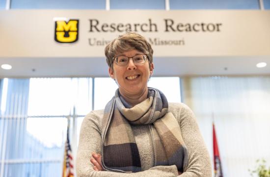 Heather Hennkens standing in front of blurred research reactor sign, wearing a gray long sleeve and plaid scarf